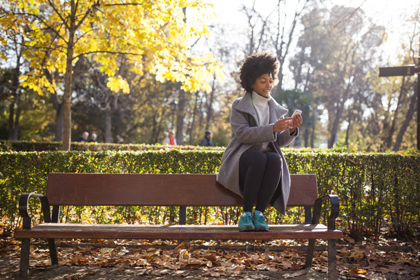 Eğleniyor güzel afro kadın parkta onun akıllı telefon ile iletişim - Fotoğraf, Görsel