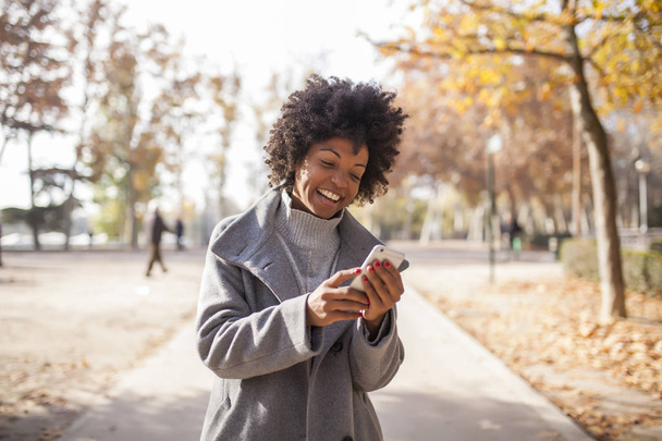 όμορφη γυναίκα Αφρο διασκεδάζοντας επικοινωνεί με το έξυπνο τηλέφωνο στο πάρκο - Φωτογραφία, εικόνα
