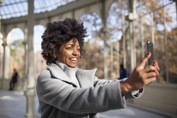όμορφη γυναίκα Αφρο διασκεδάζοντας επικοινωνεί με το έξυπνο τηλέφωνο στο πάρκο - Φωτογραφία, εικόνα