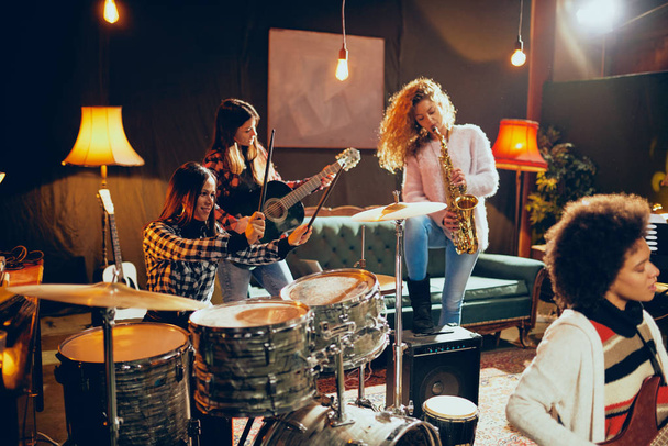 Κορίτσια που παίζουν τζαζ μουσική. Στο προσκήνιο μία γυναίκα παίζει μπάσο κιθάρα και στο φόντο άλλα τρία παίζουν ακουστική κιθάρα, σαξόφωνο και τύμπανα. Εσωτερικό στούντιο στο σπίτι. - Φωτογραφία, εικόνα