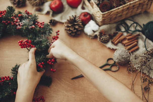Hands making rustic christmas wreath with fir branches, red berries,pine cones,rope, scissors, cinnamon, cotton on rustic wooden background. Atmospheric moody image at holiday workshop - Fotoğraf, Görsel