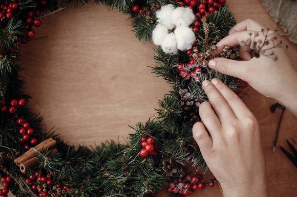 Eller, rustik Noel çelenk yapma otlar köknar şubelerinde tutan rustik ahşap arka plan üzerinde kırmızı meyveler, çam kozalakları, pamuk. Kış tatil Atölyesi'nde atmosferik moody görüntü - Fotoğraf, Görsel