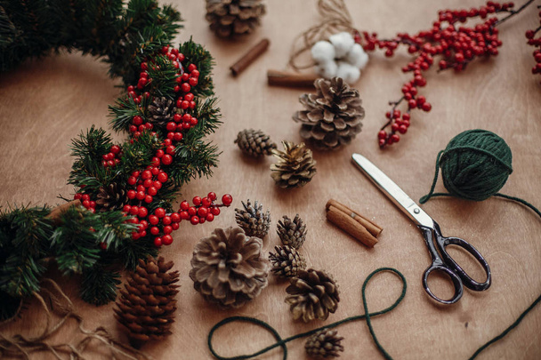 Романтичний різдвяний вінок. Ялинові гілки, вінок, червоні ягоди, соснові шишки, нитки, ножиці, кориця, бавовна на сільському дерев'яному тлі. Робимо різдвяний вінок на
 - Фото, зображення