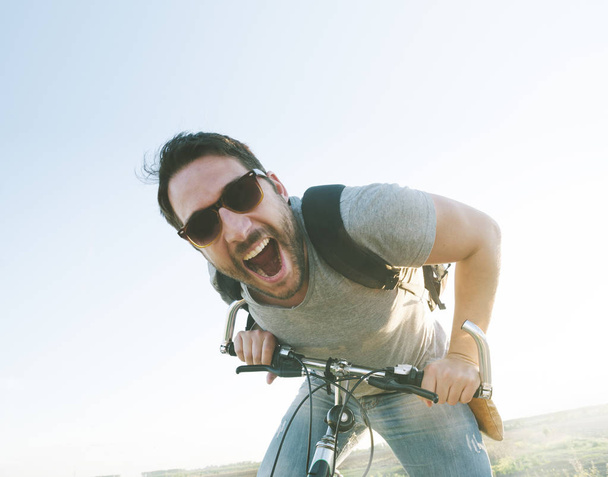 Enthusiastic hipster guy with retro sunglasses having outdoor fun and riding a bicycle. Active sport man with excited face expression exploring and traveling by mountain bike on the road. Vintage film filter style image. - Photo, Image
