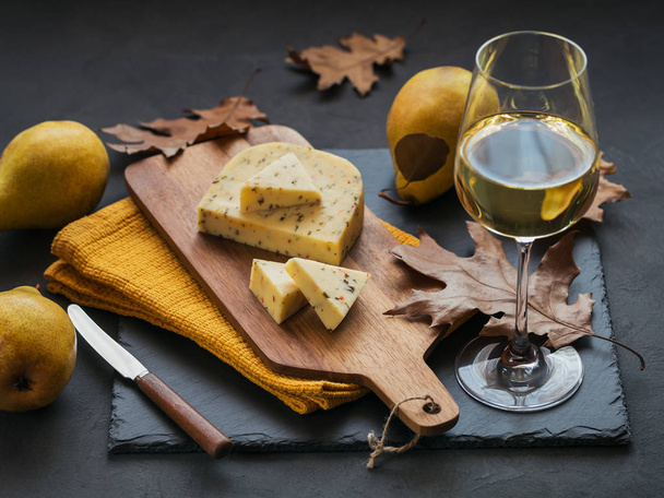 A glass of white wine was served with cheese in a cutting board on dark background. Autumn picnic with cheese, wine and dry leaves in rustic style. - Photo, Image