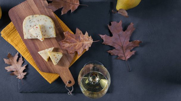 A glass of white wine was served with cheese in a cutting board on dark background. Autumn picnic with cheese, wine and dry leaves in rustic style. Top view - Photo, Image
