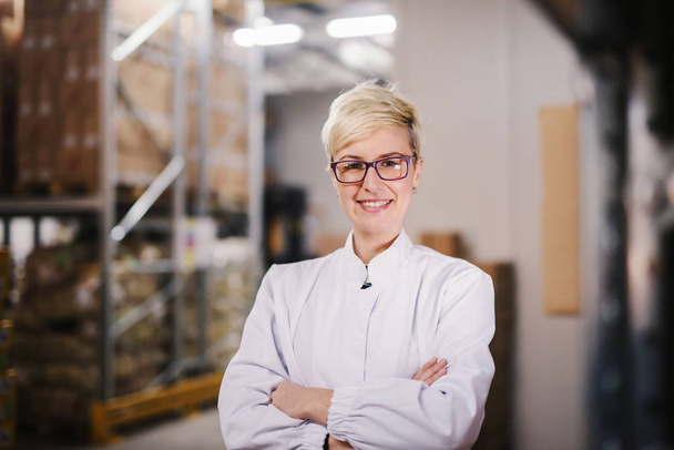 Happy Caucasian female worker posing in warehouse. Arms crossed. In background boxes on shelves. - Foto, imagen