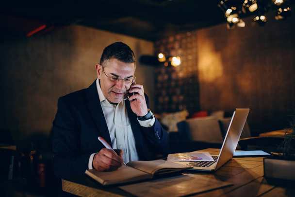 Hombre de negocios de mediana edad que usa el teléfono inteligente para llamadas de negocios y escribir en la agenda mientras está sentado en la cafetería por la noche. En el portátil de mesa, gráfico y tableta
. - Foto, imagen