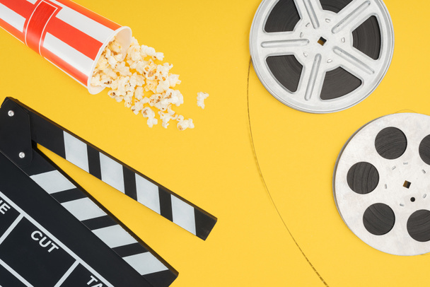 top view of clapperboard, overturned striped bucket with popcorn and film reels with cinema tape isolated on yellow - Photo, Image
