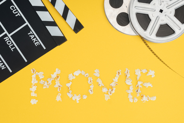 clapperboard, film reels and "movie" lettering made with popcorn isolated on yellow - Photo, Image