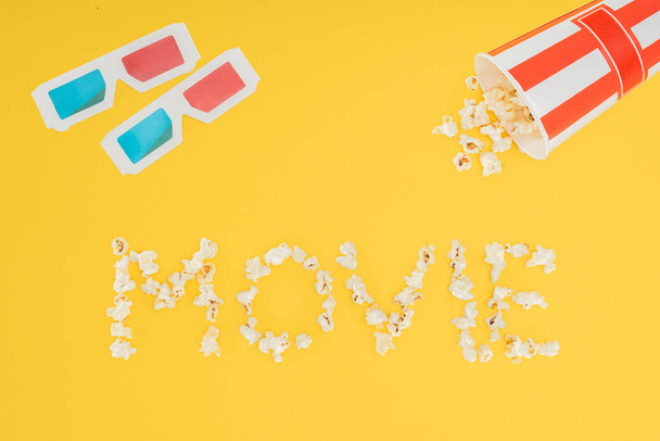 3d glasses, overturned striped bucket and "movie" lettering made with popcorn  isolated on yellow - Photo, Image