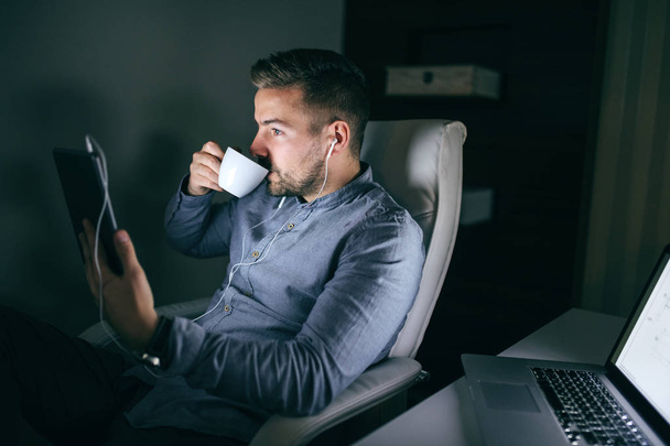 Uomo d'affari caucasico fare una chiamata su tablet e bere caffè mentre seduto in ufficio a tarda notte
. - Foto, immagini