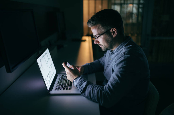 junger kaukasischer Geschäftsmann mit Brille, der sein Smartphone zum Schreiben oder Lesen von Nachrichten benutzt, während er spät in der Nacht im Büro sitzt. vor seinem Laptop. Seitenansicht. - Foto, Bild