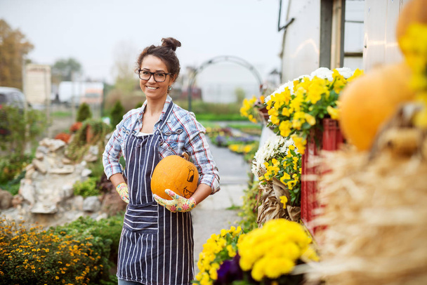 Happy female florist standing outdoors and holding pumpkin. All around potted flowers. - Photo, Image
