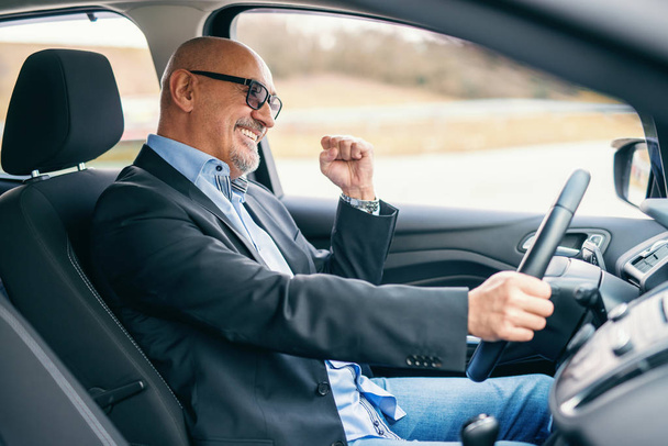 Γενειοφόρος χαμογελώντας ανώτερος ενηλίκων επιχειρηματίας οδήγηση αυτοκινήτου κατά τη διάρκεια της ημέρας. Το χέρι στο τιμόνι. - Φωτογραφία, εικόνα