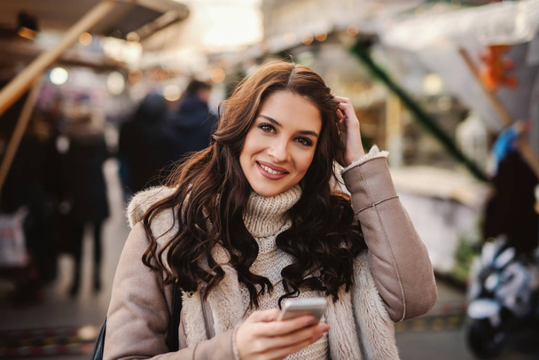 Bella donna caucasica con lunghi capelli castani in piedi sulla strada sul freddo, tenendo lo smart phone e guardando la fotocamera
. - Foto, immagini