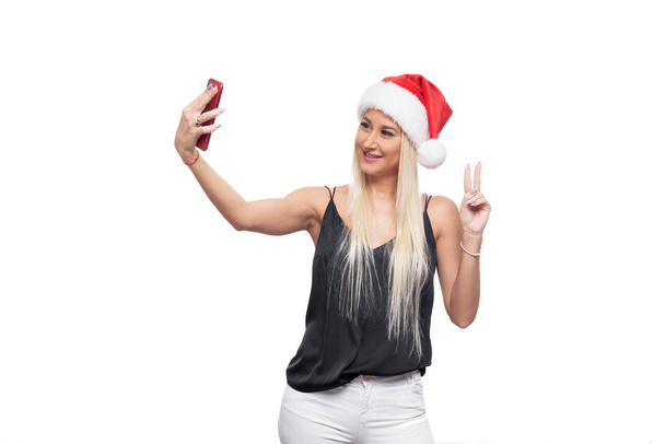 Smiling happy blond woman in black blouse and christmas santa hat make selfie on red smartphone camera over isolated white background. Sale concept - Φωτογραφία, εικόνα