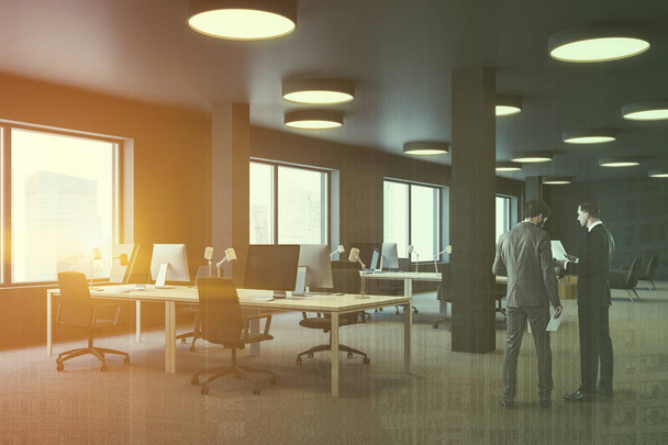 Mannen in de hoek van de industriële stijl kantoor met bruine muren, tapijt op de vloer, kolommen en rijen van witte computer tabellen met bruin stoelen. Zithoek in de achtergrond. Getinte dubbele beeldbelichting - Foto, afbeelding