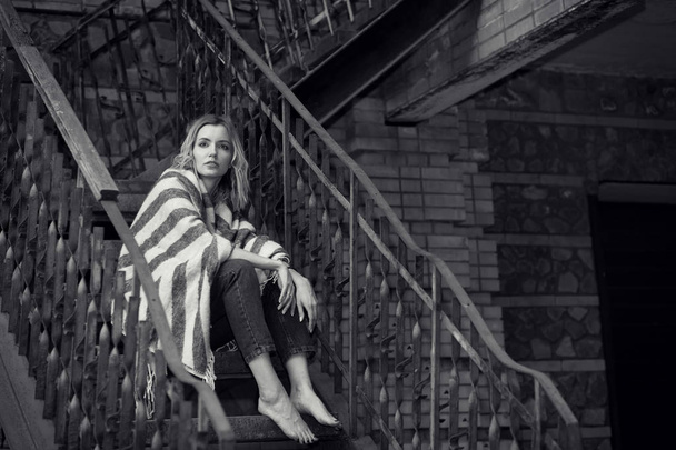 Black and white photo, portrait of a young sad woman sitting on a stone fence near the house. Depressed. - Foto, Bild