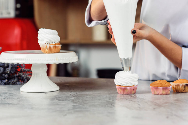 Close up hands of the female chef with confectionery bag squeezing cream on cupcakes. - Photo, image