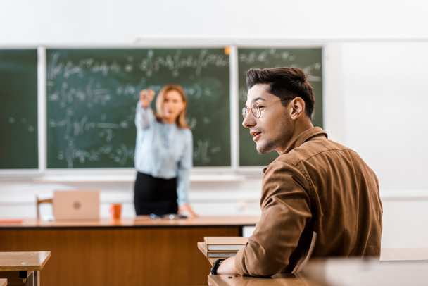 Selective focus of young student sitting in classroom with teacher on background  - Photo, Image