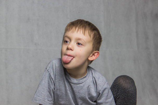 Το αγόρι με τα γκρι T-shirt κάθεται και δείχνει τη γλώσσα του - Φωτογραφία, εικόνα