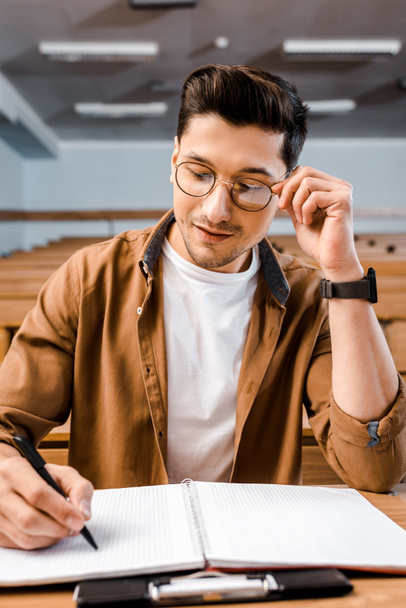 skoncentrowany studentowi w okularach siedzący przy biurku i pisanie w Notatniku podczas lekcji w klasie - Zdjęcie, obraz