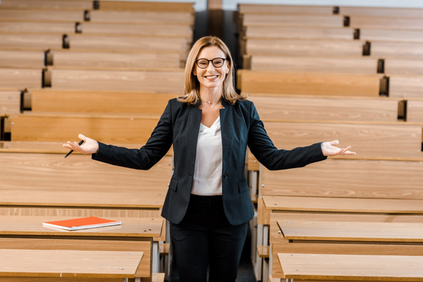 happy female university professor with outstretched hands looking at camera in classroom - Photo, Image