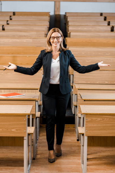 smiling female university professor with outstretched hands looking at camera in classroom - Foto, Bild