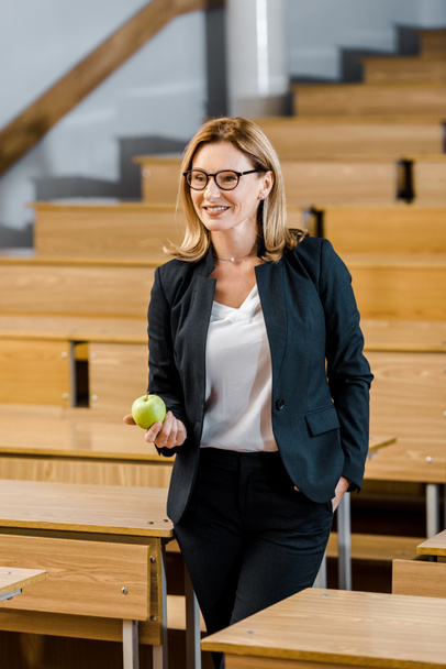усміхнена вчителька в офіційному одязі тримає яблуко в класі
 - Фото, зображення