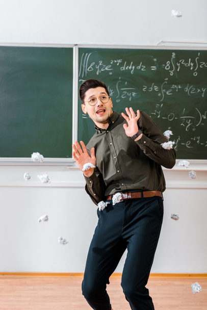 crumpled paper balls flying at frightened male teacher in classroom with chalkboard on background - Foto, Imagen
