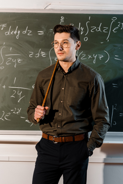 ernsthafter männlicher Lehrer in offizieller Kleidung, der einen hölzernen Zeiger vor eine Tafel mit Gleichungen hält  - Foto, Bild