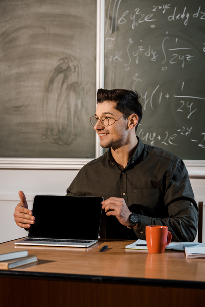 Lächelnder männlicher Lehrer mit Brille sitzt am Schreibtisch und zeigt Laptop mit leerem Bildschirm im Klassenzimmer - Foto, Bild