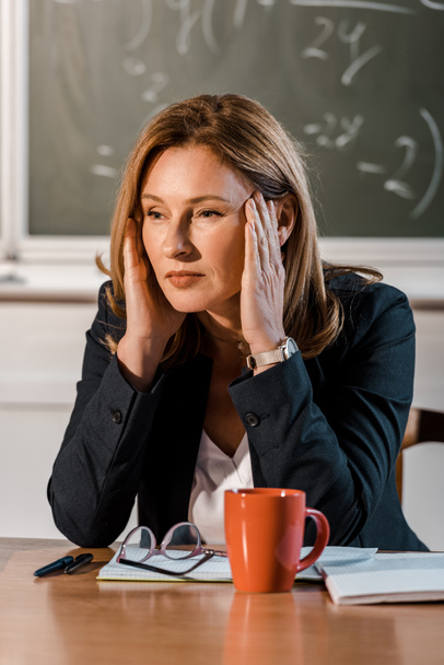 professora sentada na mesa com as mãos na cabeça e com dor de cabeça na sala de aula
 - Foto, Imagem