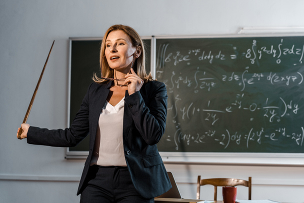 жінка-вчителька з дерев'яним вказівником тримає окуляри і пояснює математичні рівняння в класі
 - Фото, зображення