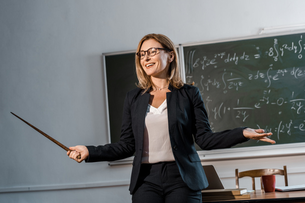 усміхнена вчителька-жіночка з дерев'яним вказівником пояснює математичні рівняння в класі
 - Фото, зображення