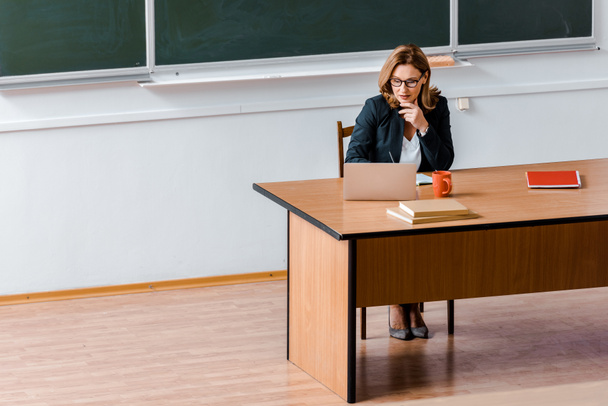 Καθηγητής Πανεπιστημίου θηλυκό σε γυαλιά, κάθεται στο γραφείο και να χρησιμοποιούν φορητό υπολογιστή στην τάξη - Φωτογραφία, εικόνα