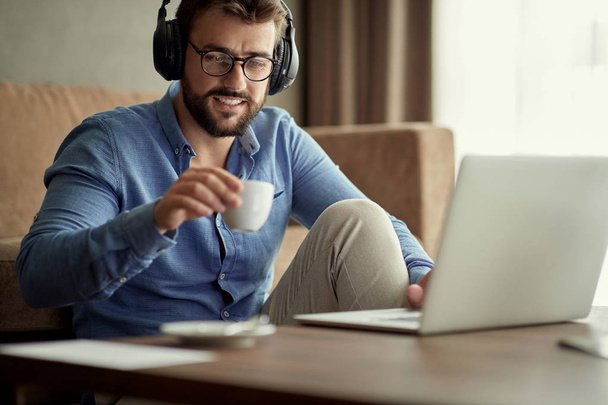 Νεαρός άνδρας με τα ακουστικά χρησιμοποιούν φορητό υπολογιστή και να πίνει καφέ στο σπίτι του - Φωτογραφία, εικόνα