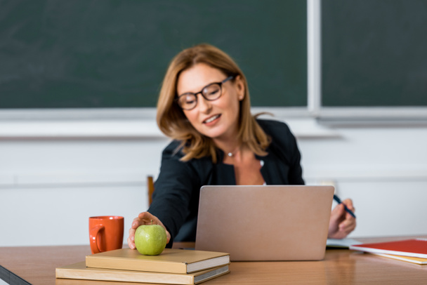 vrouwelijke leraar zit op computerbureau en bereiken voor apple in klas - Foto, afbeelding