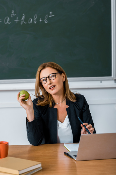 female teacher in glasses sitting at computer desk and holding apple in classroom - Foto, afbeelding