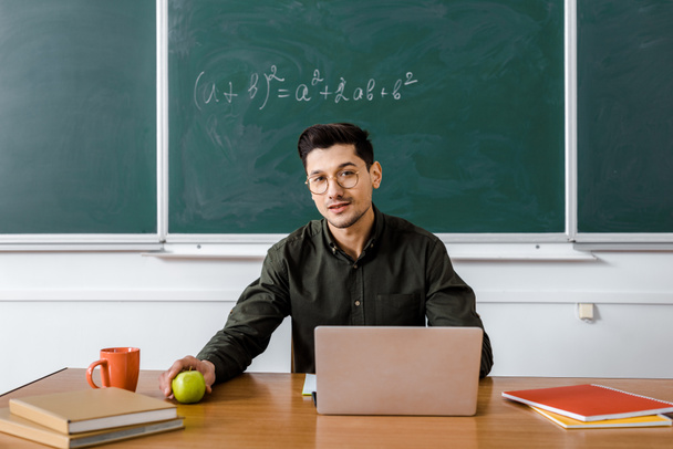 mężczyzna nauczyciel w okularach, siedząc przy biurku komputer, przytrzymując apple i patrząc na kamery w klasie - Zdjęcie, obraz