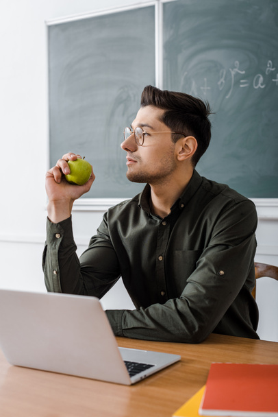 focused male teacher in glasses sitting at computer desk and holding apple in classroom - Foto, immagini
