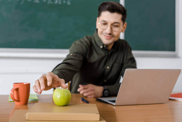 male teacher in glasses sitting at computer desk and reaching for apple in classroom - Photo, Image
