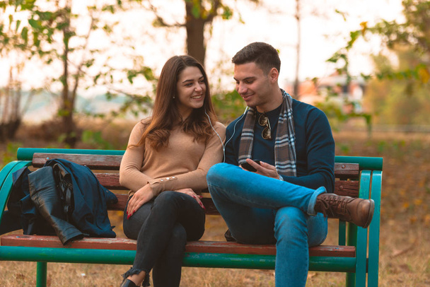 Молодая пара слушает музыку, улыбается, смотрит на телефон и сидит на скамейке в парке
. - Фото, изображение