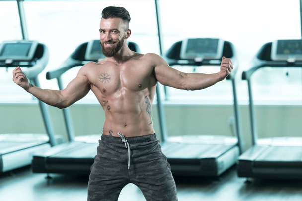 Hombre joven guapo de pie fuerte en el gimnasio y músculos de flexión - Muscular atlético culturista modelo de fitness posando después de los ejercicios - Foto, Imagen