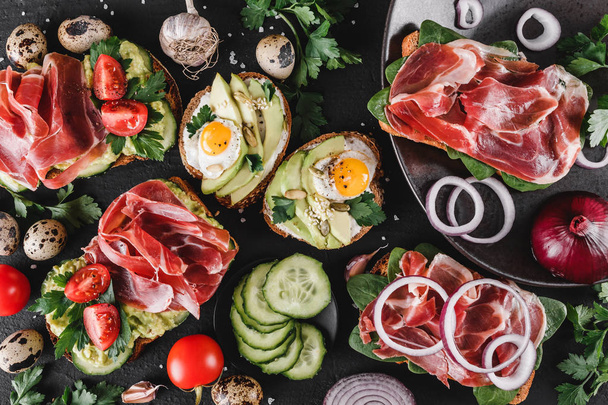 Sandwiches and bruschetta with prosciutto, fried quail egg, avocado, cucumber, tomatoes, spices and greens on black stone background. Clean eating, healthy breakfast. Top view, flat lay - Photo, Image