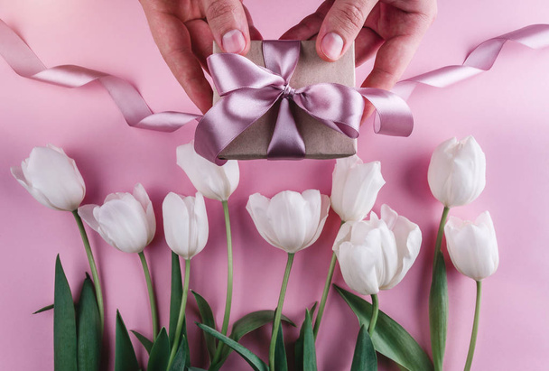 Male hands holding a gift with ribbons over pink background with white tulips. Greeting card or wedding invitation. Flat lay, top view, copy space - Photo, Image