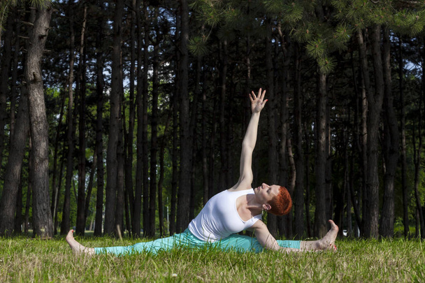 Exercise in nature-Redhead girl doing Yoga in the park, pine trees behind, sunlit, horizontal - Foto, Imagen