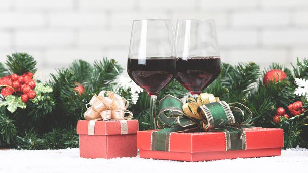 Two red wine glasses set next to red gift boxex on snowy table prepare for celabraion, decorate with pine branches and holly berries. Blurred white brick wll copy space at top part. - Фото, зображення