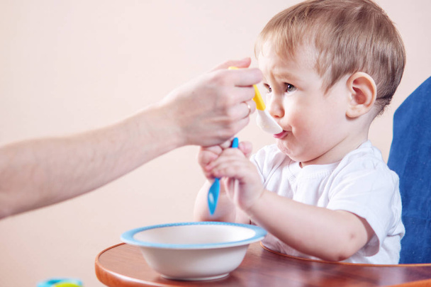Маленький милый мальчик ест на стуле на кухне. Мама кормит, держа в руках ложку каши
 - Фото, изображение